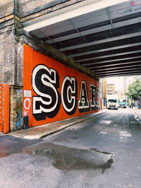 A street scene under a railway overpass in Shoreditch, N1, showing a vibrant orange wall featuring bold graffiti-style lettering spelling 'SCAR'. The pavement beneath displays a wet patch and some dirt, indicating recent cleaning or weather effects. In the background, a white van and other vehicles are seen driving along the road, with a few trees and buildings lining the street. The lighting is natural, highlighting the contrast between the colorful wall and the urban environment. The scene reflects urban aesthetic and street art, with no visible cleaning tools or surfaces, but relates to surface cleaning and maintenance in a city context, aligning with Shoreditch Carpet Cleaning's focus on deep cleaning and surface hygiene for residential and commercial spaces.