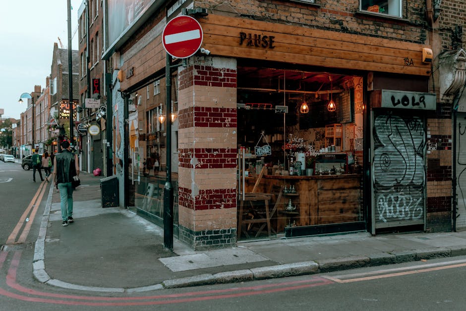 The exterior of a brick-built commercial property on a city street corner, featuring a glass-fronted shop with a wooden sign that reads 'PAUSE'. The shop has a partially open entrance with warm indoor lighting visible through the large glass window. The sidewalk in front is clean with a curb separating it from the road, which has double yellow lines along the edge. Nearby, there are pedestrians walking, streetlights, and various buildings in the background. The setting appears to be an urban area in Shoreditch, with a focus on the well-maintained exterior that reflects a clean, inviting space, consistent with professional cleaning standards by Shoreditch Carpet Cleaning for surface cleaning and maintenance purposes.