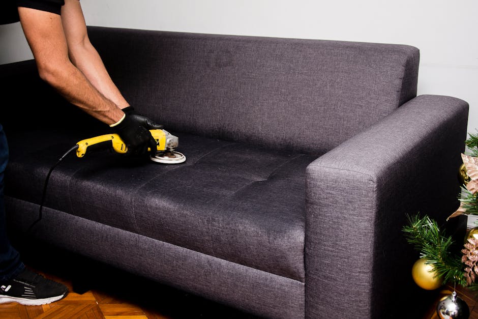 A person in black gloves is using a handheld electric grout and fabric cleaning tool on a dark grey fabric sofa in a living room. The sofa has a modern design with a high backrest and armrests, situated on a wooden floor. To the right of the sofa, there is a decorated Christmas tree with gold and pink ornaments, and in the background, a white wall is visible, illuminated by natural or soft indoor lighting. The surface of the sofa appears clean and well-maintained, with a focus on deep cleaning and sanitation as part of overall domestic cleaning services provided by Shoreditch Carpet Cleaning.