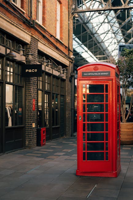 A traditional red British telephone booth situated on a paved pedestrian street in Shoreditch, London. The booth stands next to a brick building housing Shoreditch Carpet Cleaning's shopfront, with large glass windows and dark metal framing. The building's exterior features decorative brickwork and signage for P&C. Overhead, a glass and metal canopy structure extends across the street, allowing natural light to illuminate the area. The pavement is clean with a smooth finish, highlighting the urban environment and the vibrant, iconic telephone kiosk as a focal point. This scene embodies a blend of classic London architecture and modern street aesthetics, ideal for showcasing surface cleaning and maintenance in a busy commercial area, consistent with the themes of deep cleaning and hygiene emphasized by Shoreditch Carpet Cleaning.
